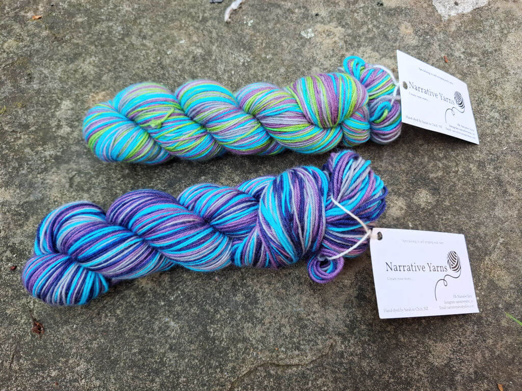 Two skeins of colourful yarn lie on a textured stone surface. The top skein has a mix of green, pink, and blue, while the bottom features purples and blues. Labels reading "Narrative Yarns" are attached.