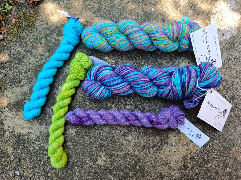 Four skeins of yarn in blue, green, purple, and multicoloured hues are arranged on a textured stone surface, creating a vibrant, cosy display.