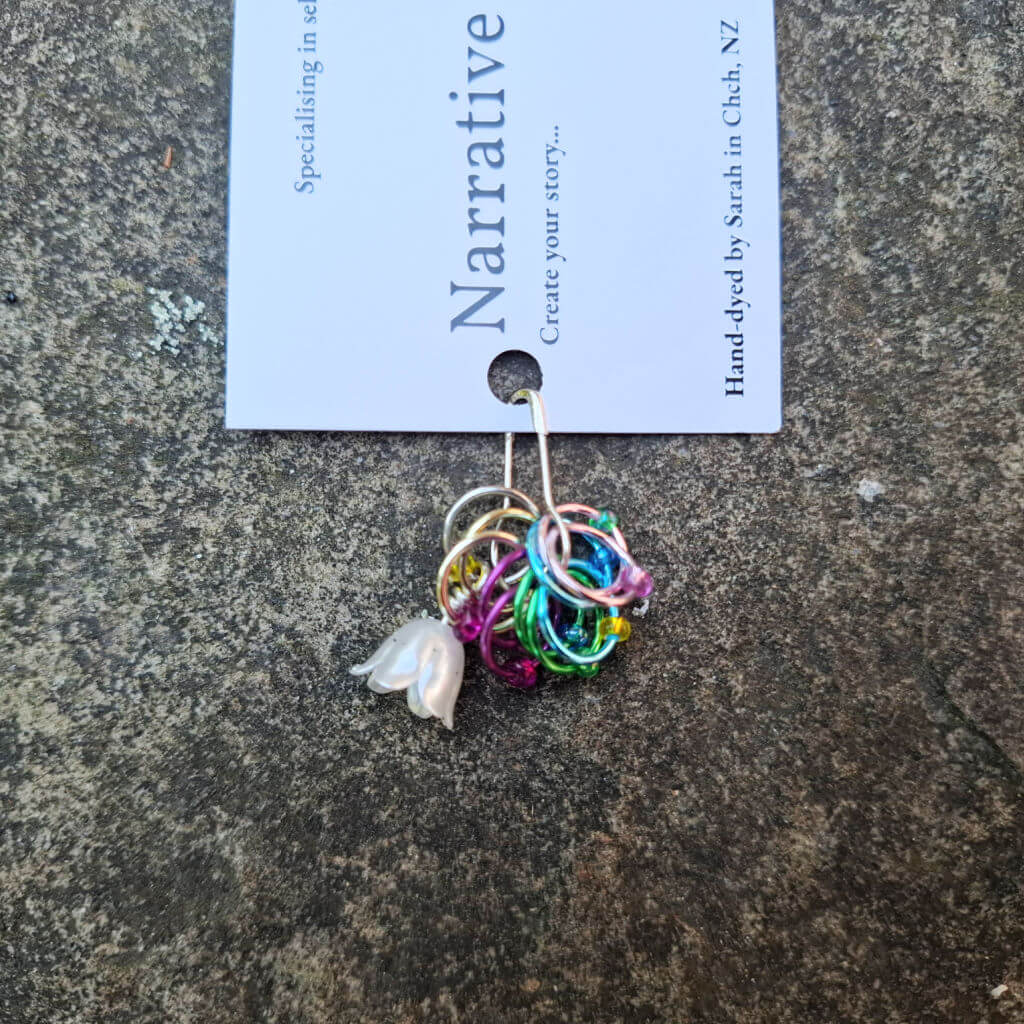 A cluster of colourful, interlinked rings with a small flower charm attached, displayed on a “Narrative Yarns” card against a textured stone background.
