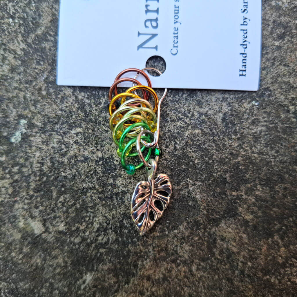 Colourful stitch markers with wire loops in shades of red, gold, green, and silver are pinned to a card. A metal leaf charm hangs below.
