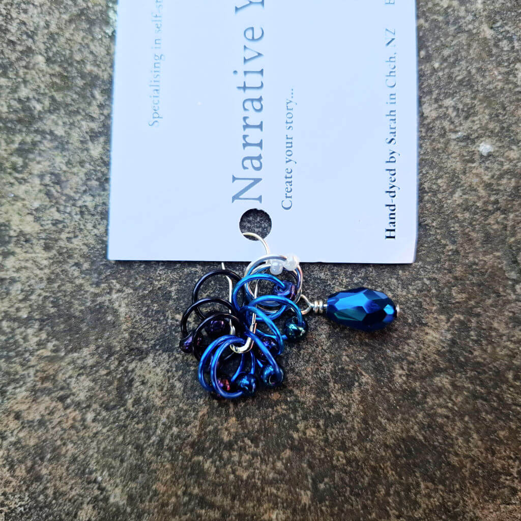 Blue and purple stitch markers with a teardrop gemstone hang on a card labeled "Narrative Yarns." They're placed on a textured stone surface.