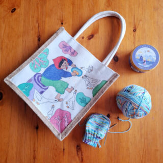 A tote bag with a colourful illustration of a woman feeding ducks, placed on a wooden table. Nearby are a ball of multicoloured yarn and a partly-knitted sock.