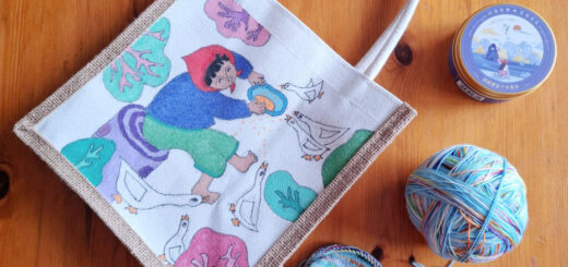 A tote bag with a colourful illustration of a woman feeding ducks, placed on a wooden table. Nearby are a ball of multicoloured yarn and a partly-knitted sock.