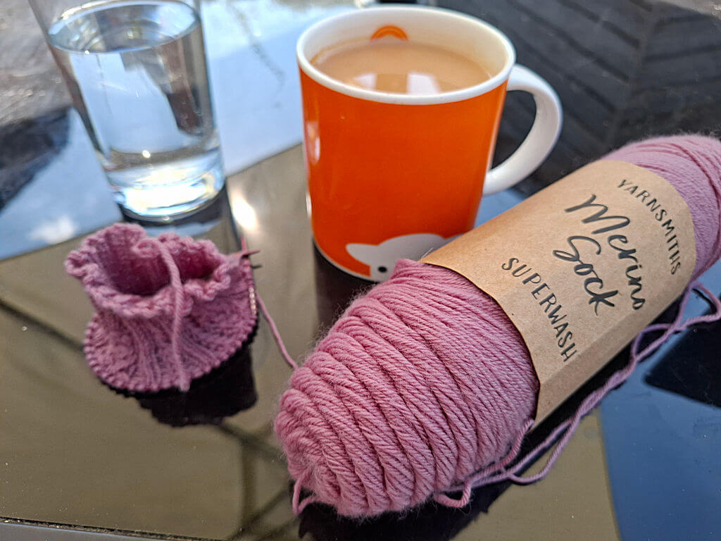 A cosy scene with pink yarn labeled "Merino Sock" and a small knitted sock beside an orange mug of coffee. A glass of water is in the background.
