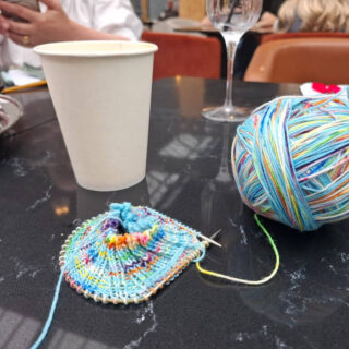 A colourful knitted sock in progress with vibrant yarn sits on a dark table near a paper cup and wine glass. A cosy, creative atmosphere.