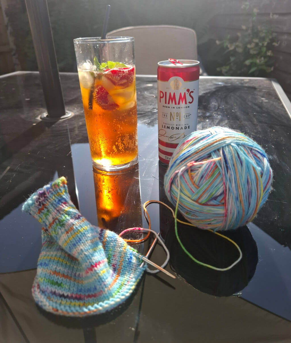 A glass of iced Pimm's with strawberries and mint sits beside a can on a glass table. Nearby, a ball of colourful yarn and a partly knitted sock are in sunlight.