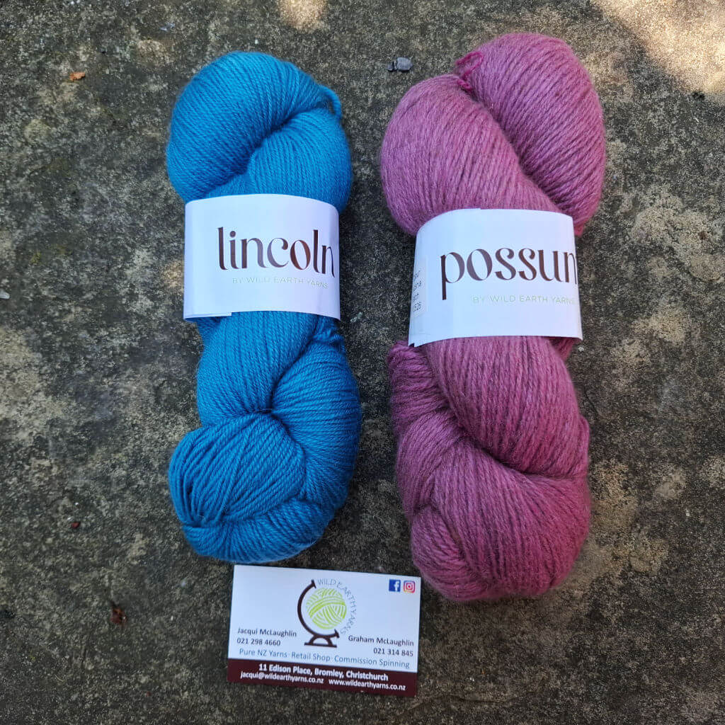 Two skeins of yarn lie on a textured gray surface. The left skein is vibrant blue and labeled "Lincoln," the right skein is pink with "Possum." A business card sits below.