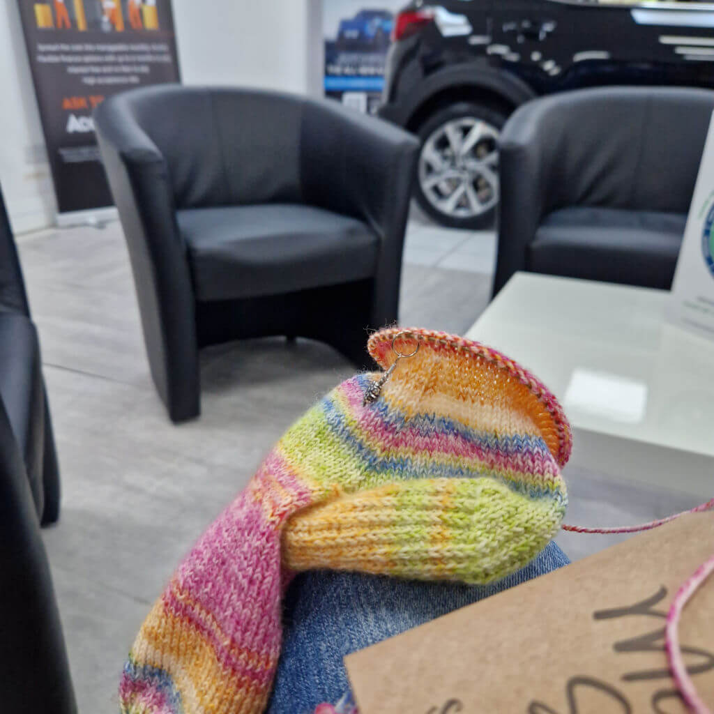 Colourful striped knitted sock rests in a car dealership waiting area, contrasting with black armchairs, a white table, and a vehicle behind.