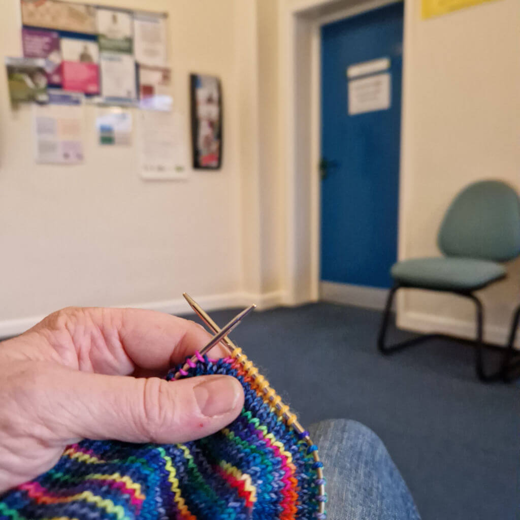 Hand knitting colourful striped sock with metal needles in a quiet waiting room, featuring a blue door, chair, and noticeboard in the background.