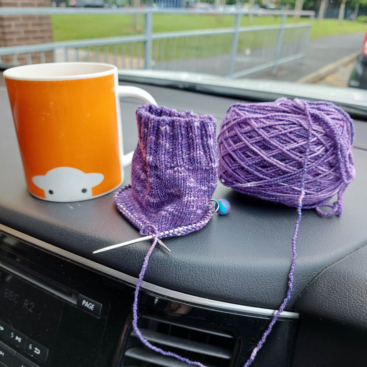 Orange mug with a sheep face, purple knitted sock-in-progress, knitting needles, and yarn on a car dashboard create a cosy crafting scene.