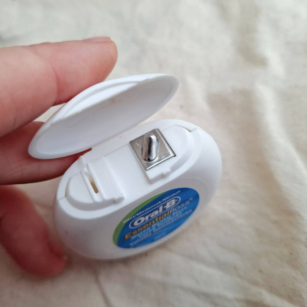 Close-up of a hand opening a white Oral-B Glide dental floss container, revealing the floss cutter, against a soft beige fabric background.