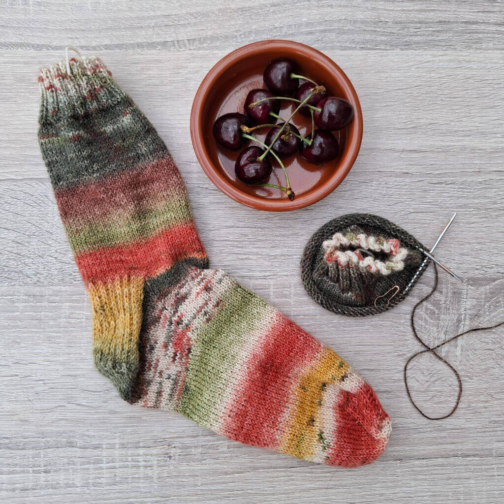 Hand-knit striped wool socks lie beside cherries in a bowl and a yarn ball with needles on a wood table, creating a cosy handmade scene.