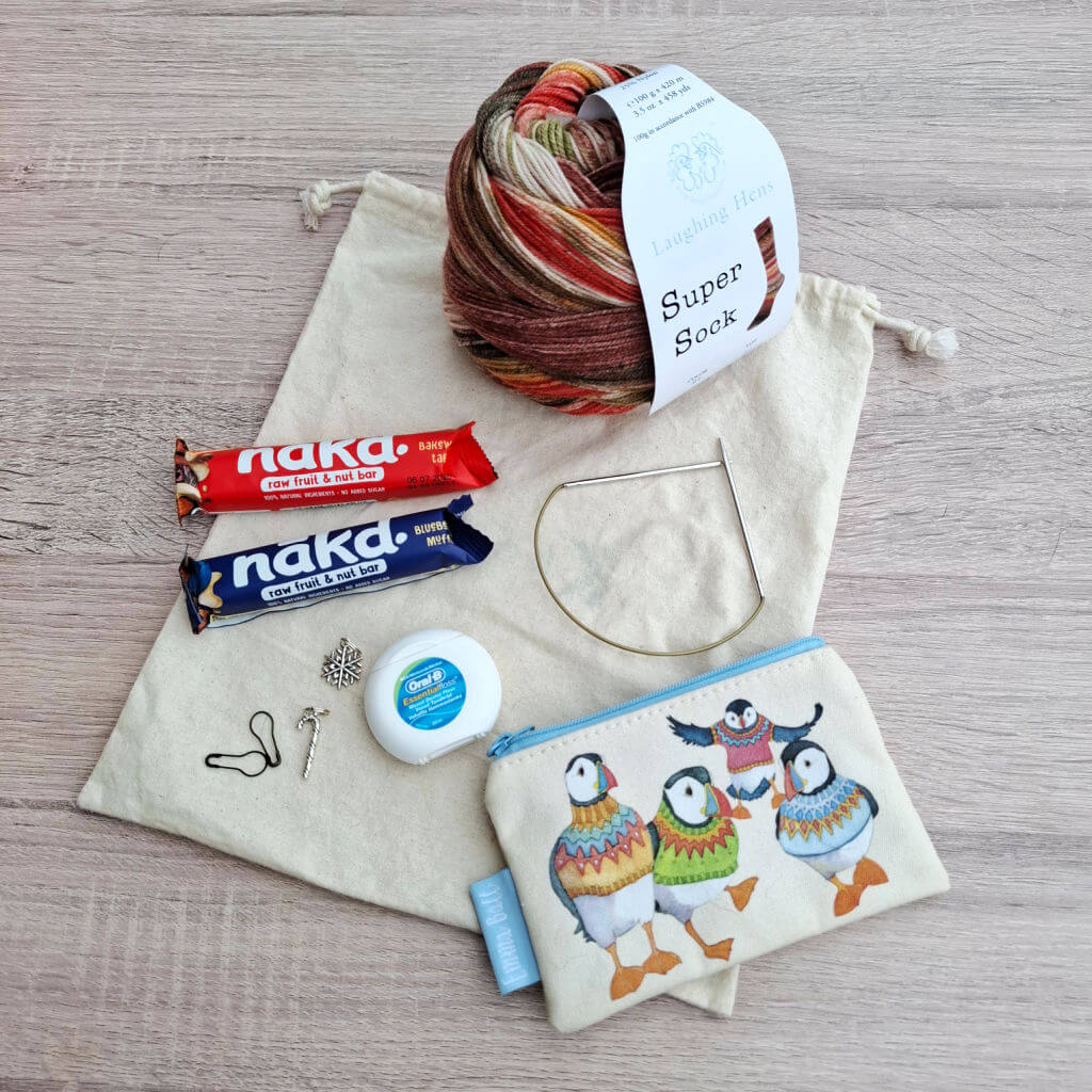 Emergency sock knitting supplies on a linen pouch: colourful yarn, Nakd fruit bars, circular knitting needle, stitch markers, dental floss, and a small zipped pouch with puffins on it.