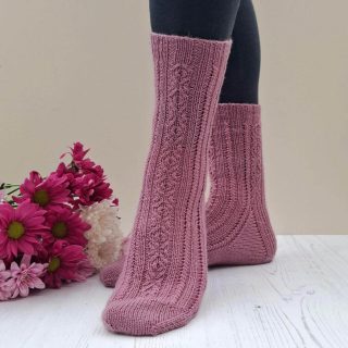 Pink twisted stitch knit socks modelled on feet beside a bouquet of magenta and white chrysanthemum flowers, creating a cosy, handcrafted mood. Winwick Mum | September Socks