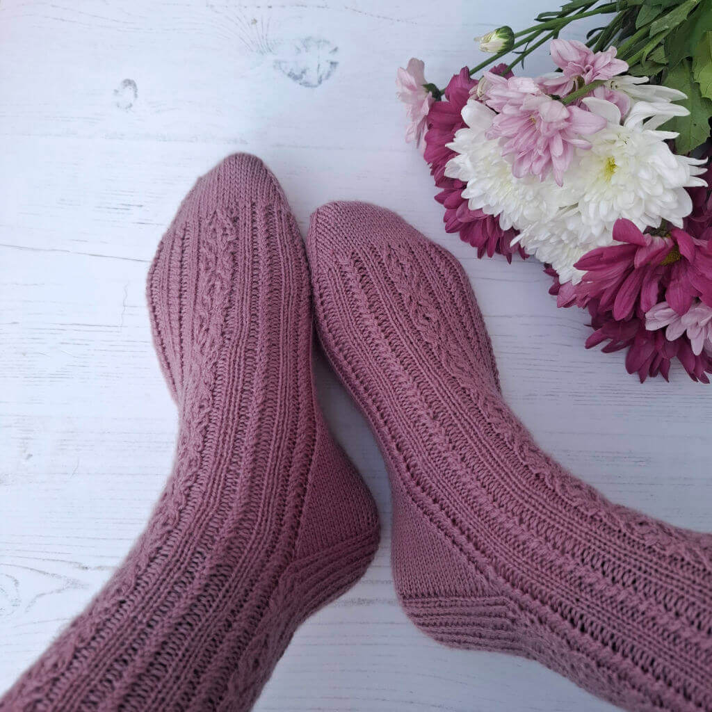 Hand-knit mauve wool socks rest on a white wooden surface beside a bouquet of pink, purple, and white chrysanthemums, creating a cosy tone.