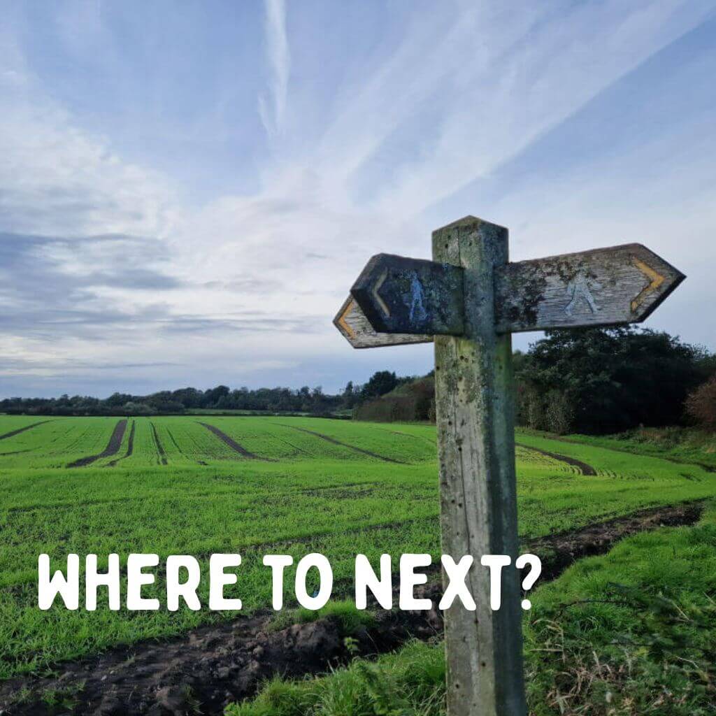 A wooden signpost in the countryside. In the background is a green field of crops and the blue sky is streaked with clouds. Text reads "Where to next?"