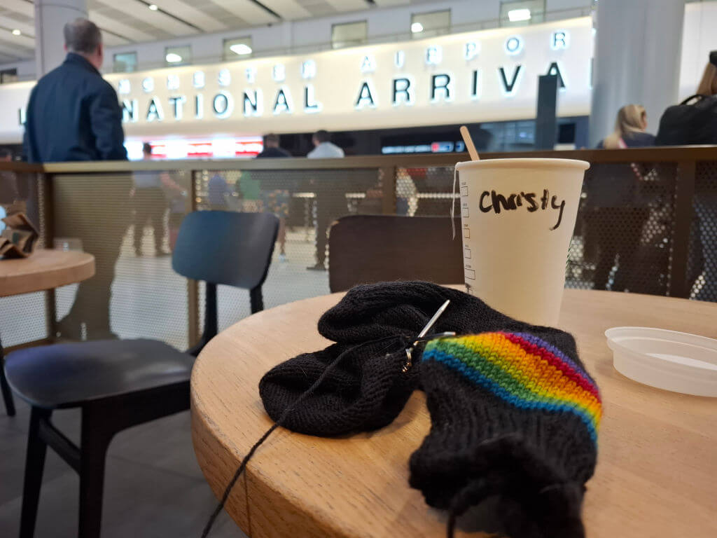 At an airport arrivals hall, a rainbow-striped knitted sock and black yarn rest on a café table beside a takeaway drink cup.