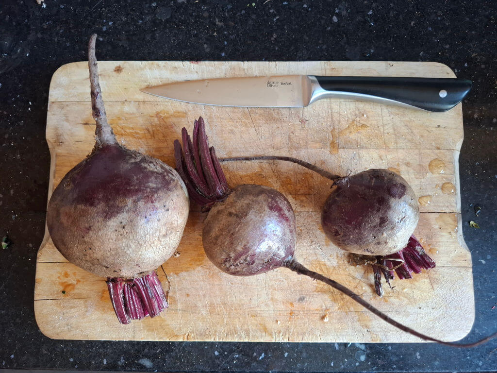 Three freshly harvested beets with leafy stems rest on a worn wooden cutting board beside a kitchen knife, creating a rustic, earthy scene.