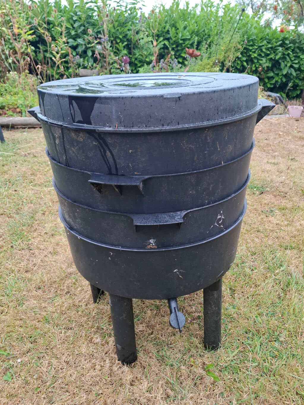 Large black plastic compost bin stands on short legs in a grassy garden, with a fitted lid, molded side handles, and lush greenery behind it.
