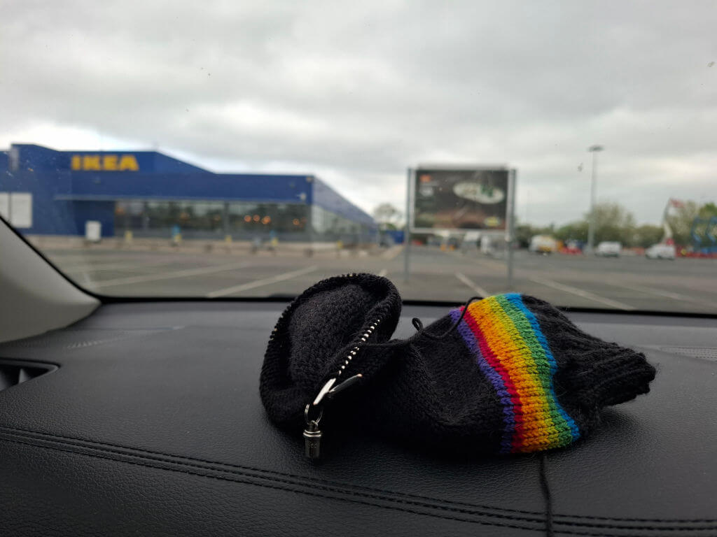 Black wool sock with a rainbow-striped band rests on a car dashboard, with an IKEA store, car park, and cloudy sky visible outside.