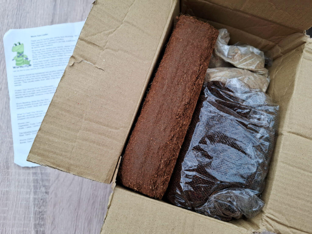 Open cardboard box containing a long coir brick and a bag of composting worms sealed in plastic, with an instruction sheet beside it.