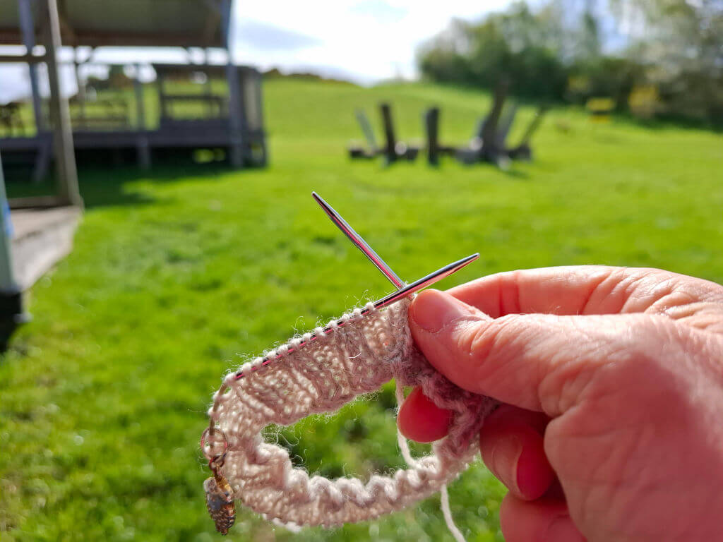 Hand holds a partially knitted pink-and-white sock cuff on knitting needles outdoors, with a sunlit green lawn and blurred outdoor eating space behind.