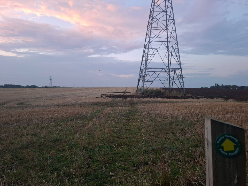 A harvested field at dusk beneath pink clouds, with a towering electricity pylon, distant poles, and a footpath marker creating a quiet rural scene.