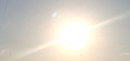 Bright sun blazes through a hazy sky with solar eclipse lens flare, silhouetted trees below, and faint power lines creating a warm, tranquil mood.
