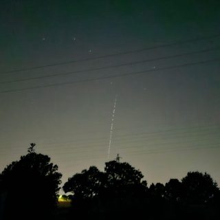 Night time landscape with silhouetted trees, power lines, and a faint vertical dotted light trail of Starlink satellites rising into the hazy sky; quiet, mysterious mood.