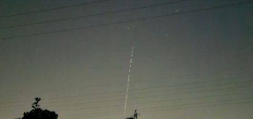 Night time landscape with silhouetted trees, power lines, and a faint vertical dotted light trail of Starlink satellites rising into the hazy sky; quiet, mysterious mood.