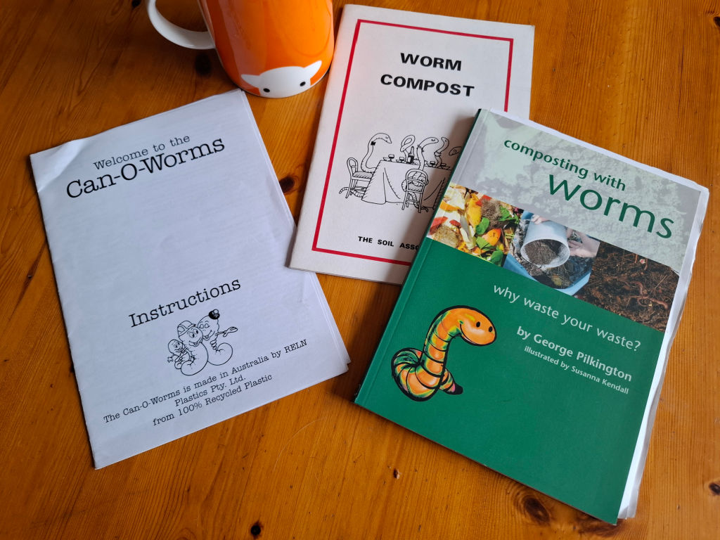 Three books about worm composting on a wooden table next to an orange mug with a sheep head on it.