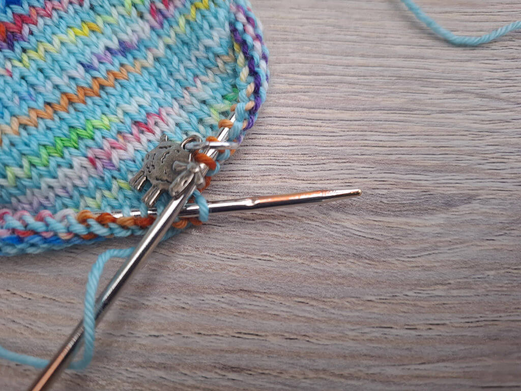 Close-up of coloruful knitted fabric on circular metal needles, with blue, pink, yellow, and green yarn on a wooden surface; cosy craft scene. The needle tip has a dent in it caused by a cat chewing the needle.