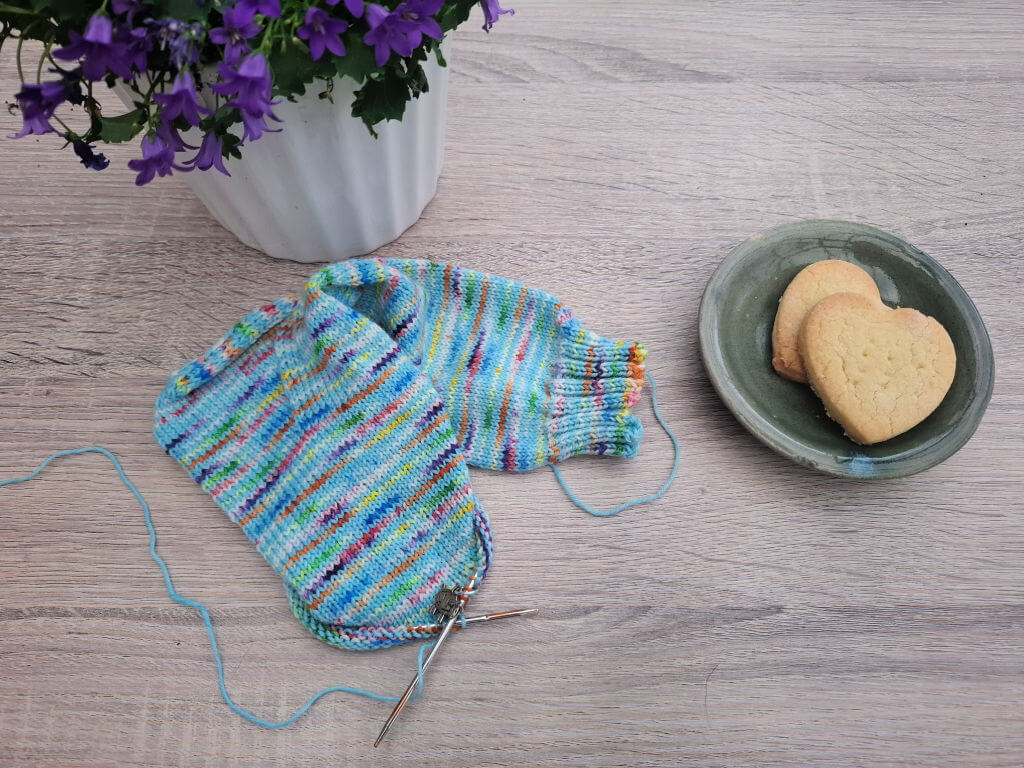 Colourful striped knitted sock in progress with needles and loose yarn, beside heart-shaped biscuits and purple flowers on a wooden table.