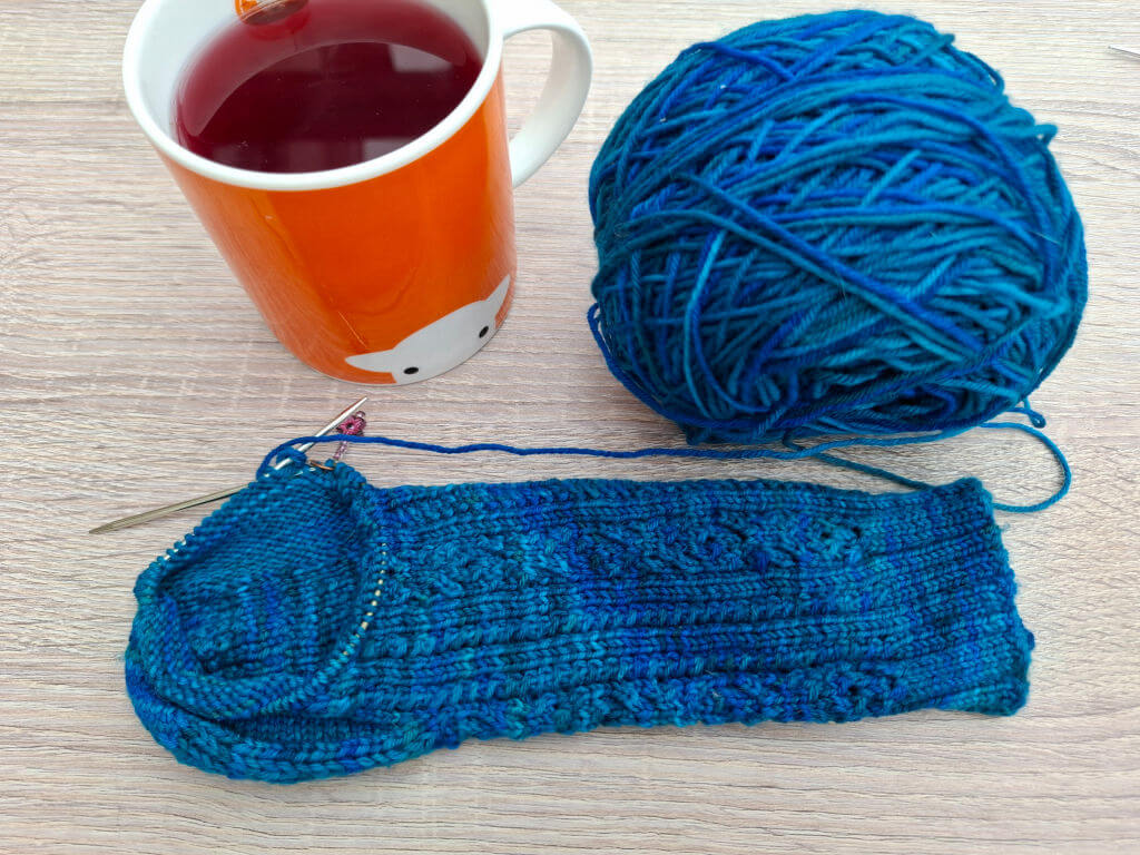 Blue yarn ball and partially knitted sock with needles beside an orange mug of tea on a light wood table, creating a cosy crafting scene.