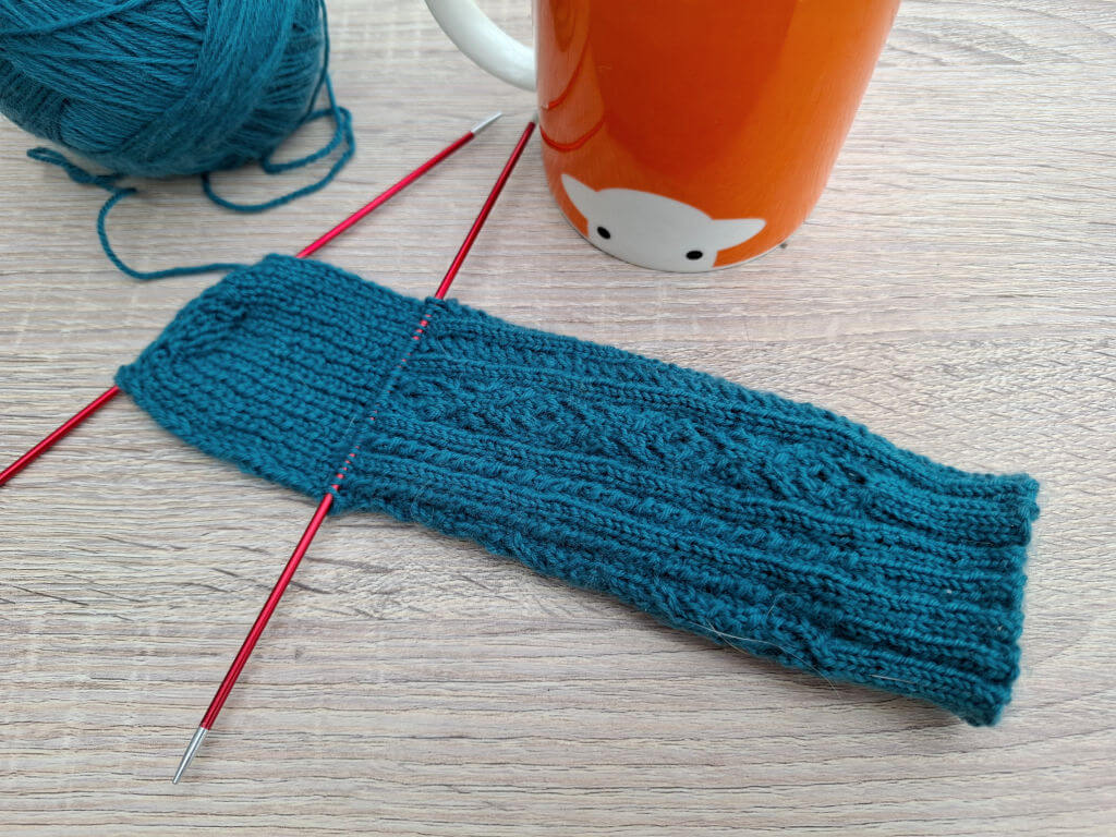 Teal knitted sock on red needles beside a matching yarn ball and orange sheep mug, creating a cosy, handmade crafting scene.