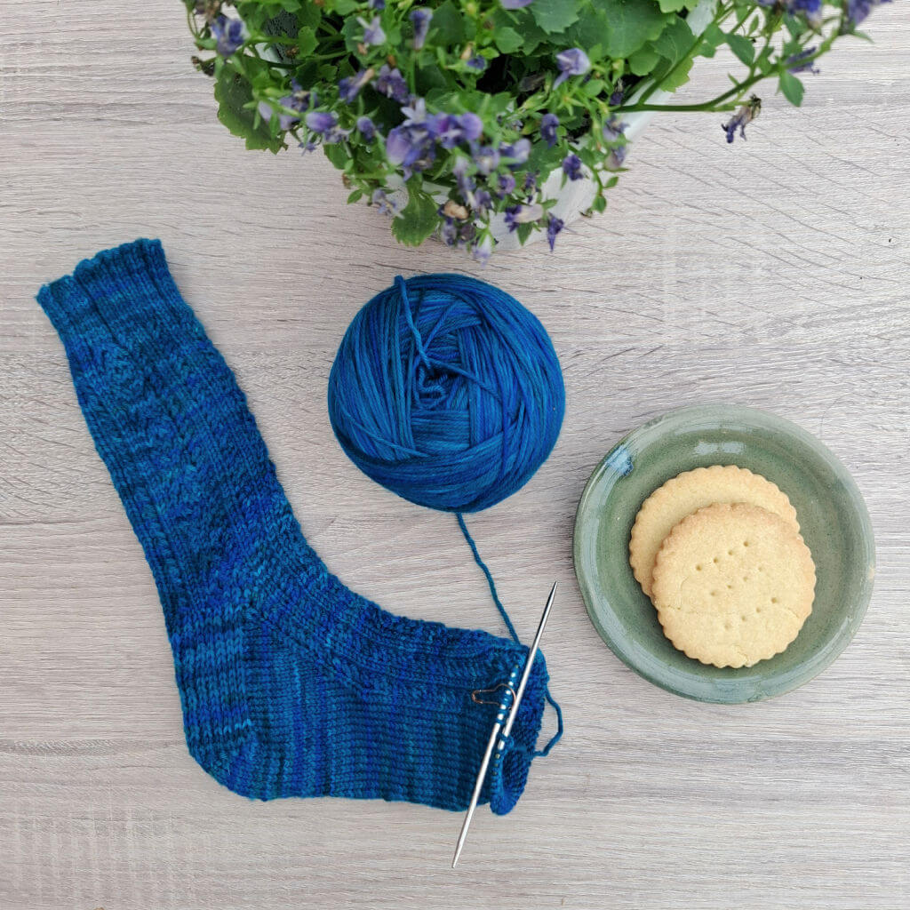 A blue patterned partly-knitted sock and a ball of yarn on a wooden table next to a green saucer with two biscuits and a pot plant with purple flowers