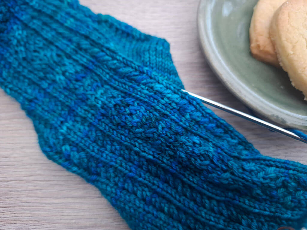 Close-up of a teal-blue knitted sock with textured stitches and metal needles beside a plate of biscuits, creating a cosy, handmade mood.