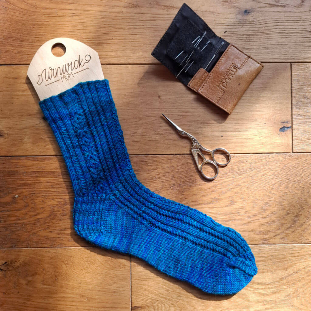 A hand-knit bright blue wool sock lies on a wooden surface beside embroidery scissors and a leather needle case, creating a warm, handmade craft scene.