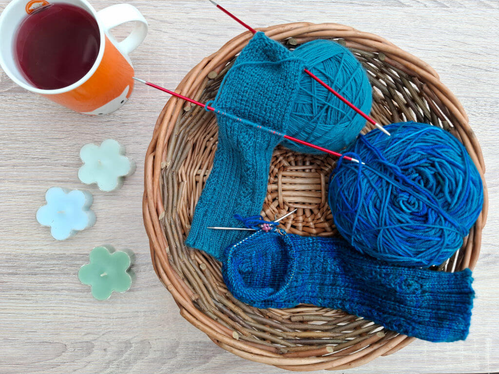 Top-down view of a wicker basket with teal and turquoise yarns, knitted socks, red needles, orange tea mug, and pastel flowers; cosy craft scene.
