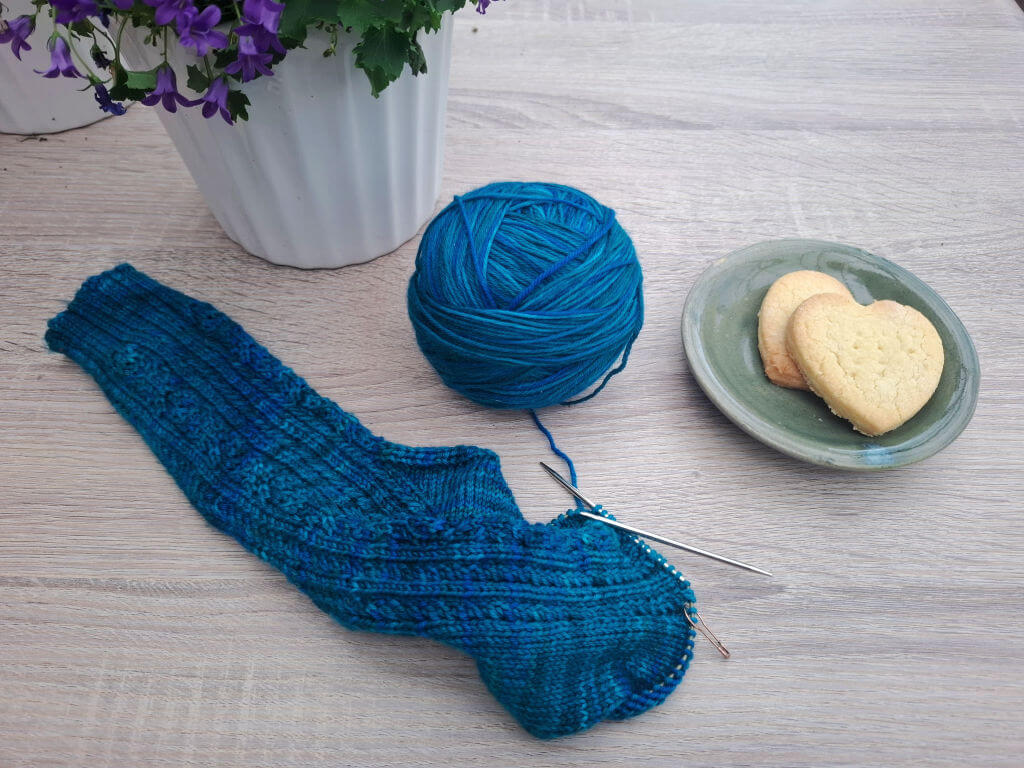 Overhead view of teal knitted sock, matching yarn ball and needles beside heart-shaped biscuits and purple flowers, creating a cosy mood.