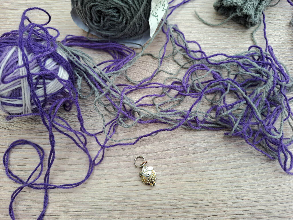 A tangled pile of purple and grey yarn sprawls across a pale wood surface beside yarn balls and a tiny gold-and-white charm, creating a messy, crafty mood.