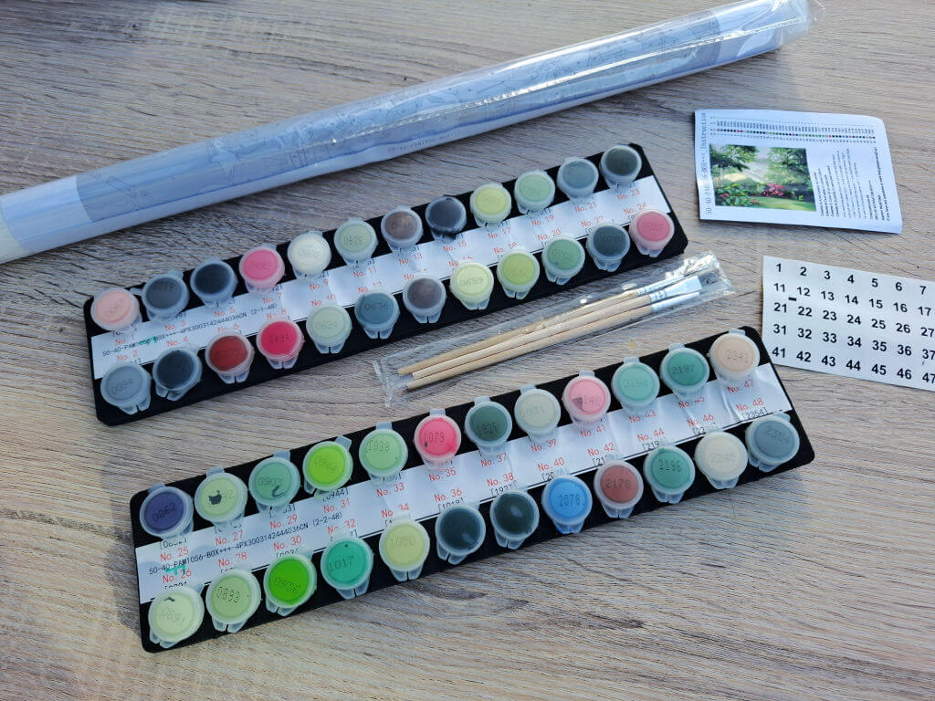 Two rows of colourful paint pots, paintbrushes, and a wrapped art tool lie on a wooden table beside an instruction card, ready for a calm craft project.