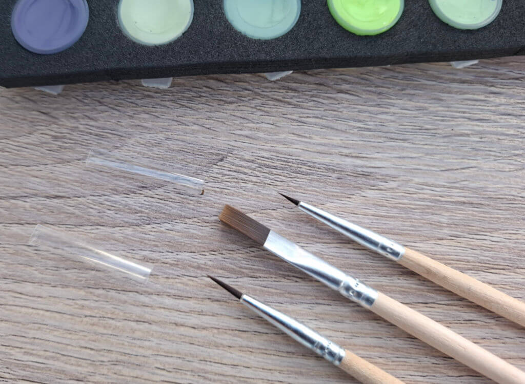 Three paintbrushes with wooden handles lie across a wood surface beneath pastel paint pots, ready for a calm art session.