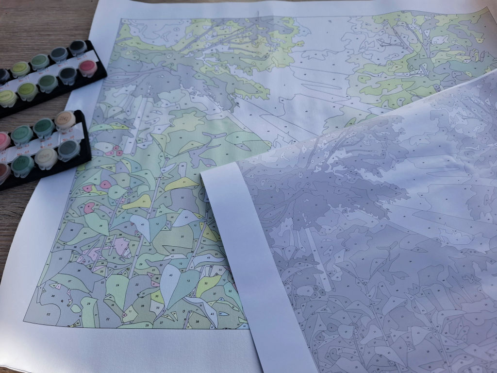 A detailed colour-by-number map rests on a table beside paint pots, with a second uncoloured sheet overlapping it, suggesting a calm painting project in progress.