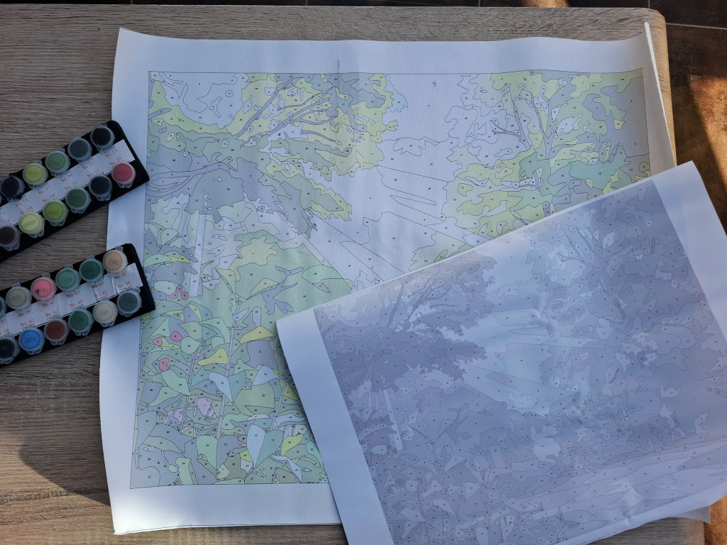 A printed paint-by-numbers canvas lies on a wooden table beside paint pots, partly covered by a paper printout sheet of the same canvas; the scene feels calm and creative.