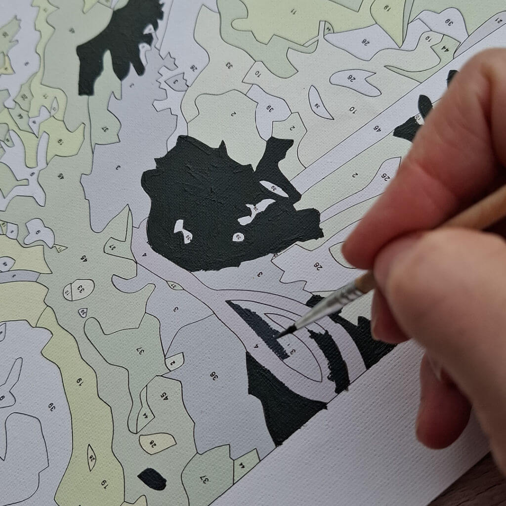 A hand uses a fine brush to paint black sections on a numbered paint-by-numbers canvas, outlining a shape with dark paint; calm, focused craft scene.