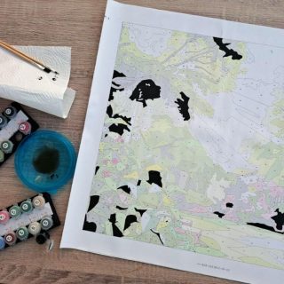 Unfinished paint-by-numbers landscape map with pastel regions and black painted shapes, surrounded by paints on a wooden desk; calm, creative mood.