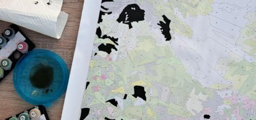 Unfinished paint-by-numbers landscape map with pastel regions and black painted shapes, surrounded by paints on a wooden desk; calm, creative mood.