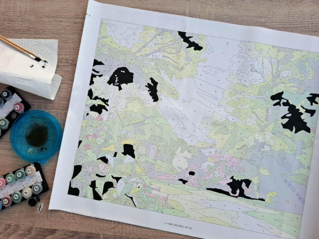Unfinished paint-by-numbers landscape map with pastel regions and black painted shapes, surrounded by paints on a wooden desk; calm, creative mood.
