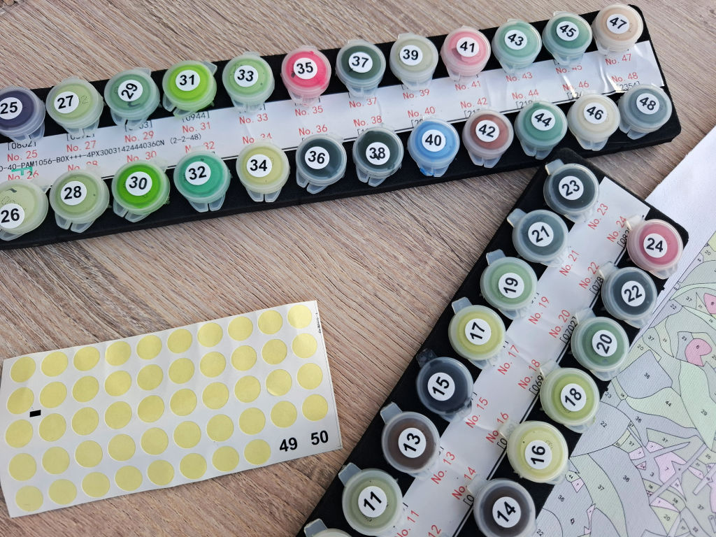 Numbered paint pots in a black tray surround a sheet of yellow stickers and a colour-by-number canvas, ready for a creative project.